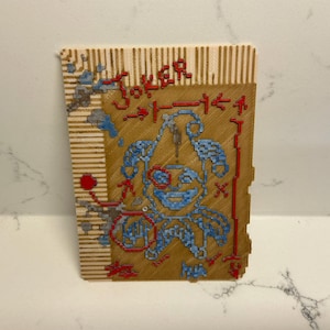 May include: A wooden playing card featuring a cross-stitch design of a joker. The card has a brown background with blue and red stitching. The word "Joker" is stitched across the top.