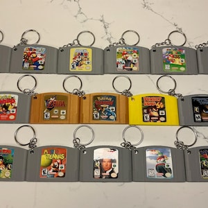 May include: A collection of sixteen Nintendo 64 game cartridge keychains arranged in three rows. Each gray, yellow, or orange plastic cartridge features a unique game label with titles like Mario Golf, Zelda, Donkey Kong 64, and Pokemon Stadium.