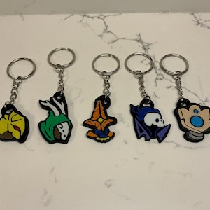 May include: Five keyrings with character designs. Each keyring has a silver ring and chain. The designs include a yellow and brown flower, a green and white character, an orange and blue design, a purple skull, and a blue and tan design.