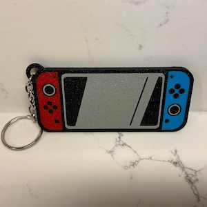 May include: A black, red, and blue keychain shaped like a video game console. The keychain has a silver key ring and chain. The console has a gray screen with a black outline and a diagonal black line.