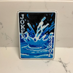 May include: A rectangular playing card featuring a blue water splash design against a black background. The word "JOKER" is vertically printed on both sides in white. The card has a white border.