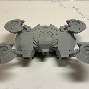 Może przedstawiać: Szary, drukowany w 3D model futurystycznego pojazdu. Model ma centralny korpus z czterema okrągłymi, obracającymi się elementami wystającymi na zewnątrz. Mały czerwony akcent jest widoczny z przodu pojazdu.