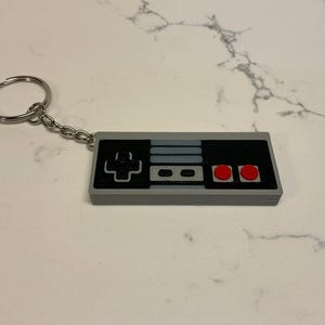 May include: A gray and black keychain shaped like a classic video game controller. The controller has red buttons and a silver key ring attached by a chain. The design is a retro gaming theme.