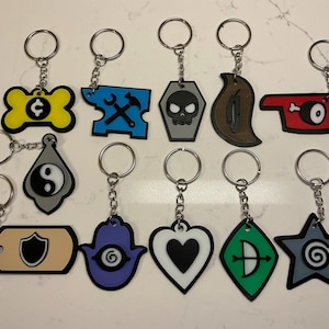 Może przedstawiać: Kolekcja breloków do kluczy z różnymi wzorami, w tym kość ze znakiem dolara, czaszka w trumnie, serce i gwiazda ze spiralą. Każdy brelok ma srebrny pierścień i łańcuszek.