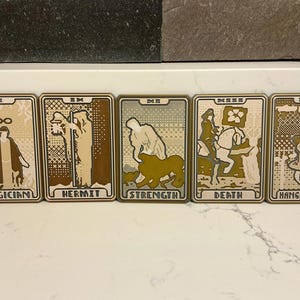 May include: Five tarot cards with brown and beige illustrations. The cards are labeled "Magician", "Hermit", "Strength", "Death", and "Hanged Man". Each card features unique imagery and a Roman numeral.