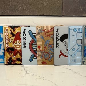 May include: A collection of playing card designs featuring the word "JOKER" and various graphics. The cards are arranged in a row, showcasing diverse artwork, including a red "RESER PARKI" sign, a smiling jester, and other unique illustrations.
