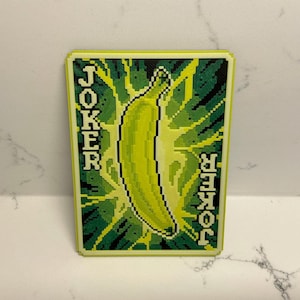 May include: A rectangular card with a pixelated design featuring a banana and the word "JOKER" on both sides. The banana is yellow, surrounded by a green and yellow lightning-like pattern, set against a black background.