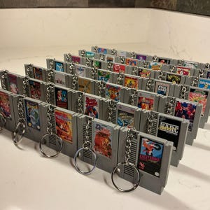 Może przedstawiać: Kolekcja szarych breloków do gier wideo. Każdy brelok przedstawia miniaturową replikę klasycznej kasety do gier Nintendo Entertainment System (NES), z kolorowymi grafikami gier i srebrnym kółkiem do kluczy.