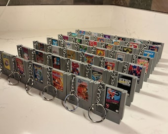 Puede incluir: Una colección de llaveros de cartuchos de videojuegos grises. Cada llavero presenta una réplica en miniatura de un cartucho de juego clásico de Nintendo Entertainment System (NES), con ilustraciones de juegos coloridas y un llavero plateado.