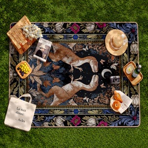May include: A rectangular picnic blanket with a black background and a floral border. Two brown and tan dinosaur illustrations are centered on the blanket. A straw hat, basket, and tote bag are on the blanket.