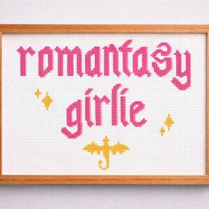 May include: Cross-stitch art in a wooden frame. The design features the words "romantasy girlie" in pink, with yellow star and dragon accents. The artwork is displayed on a white background.