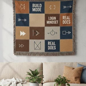May include: A patchwork wall hanging with a variety of brown, blue, and beige squares, featuring text and symbols. The text includes "BUILD MODE", "LOGIN MINDSET", and "REAL DOES". The hanging is displayed above a beige sofa in a living room.