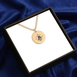 May include: Gold-colored pendant necklace with a round charm featuring a compass design and a skull wearing sunglasses. The necklace is displayed in a black box with a white interior, set against a blue background.