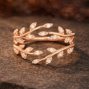 Può includere: Anello in oro rosa con un design a foglie. L'anello presenta due fasce con dettagli a forma di foglia, ciascuna incastonata con piccole gemme. Il design è evidenziato su uno sfondo neutro, sottolineando i suoi intricati dettagli.