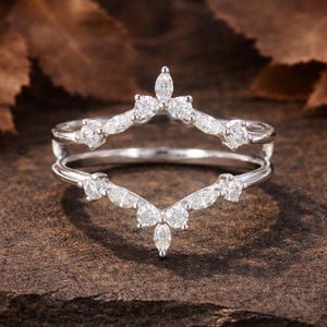 May include: A silver ring with a unique design, featuring a double band and a series of sparkling white gemstones. The gemstones are arranged in a delicate, curved pattern, creating a crown-like appearance. The ring is set against a textured, brown background.