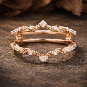 Puede incluir: Guardia de anillo de oro rosa con diseño de hojas, adornada con pequeños diamantes brillantes. El anillo presenta un diseño en forma de V, con un diamante central en la parte superior e inferior. El anillo está sobre un fondo neutro.