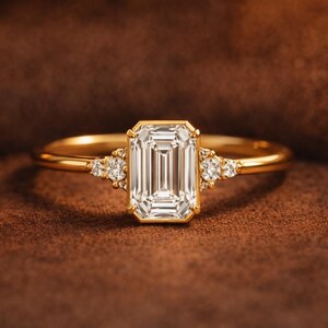 May include: A gold ring with a large, rectangular, clear gemstone as the centerpiece. Smaller, round, clear gemstones are set on either side of the main stone. The ring is set against a brown background.
