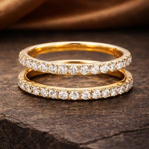 May include: Two gold rings, each set with a band of small, clear gemstones. The rings are stacked, with the gemstones sparkling. They rest on a dark, textured surface. The rings are a classic design.