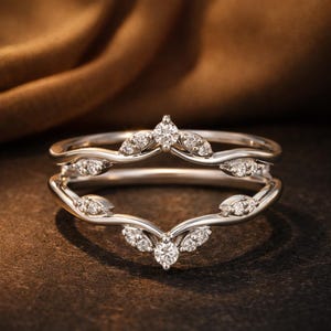 Puede incluir: Un protector de anillo plateado con un diseño en forma de hoja, adornado con pequeños diamantes brillantes. El anillo presenta un diseño curvo y abierto, perfecto para realzar un anillo de compromiso solitario. El anillo está sobre un fondo oscuro.