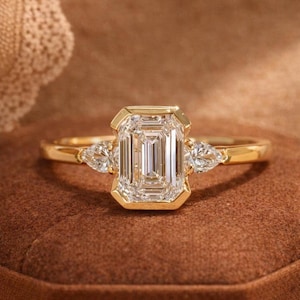 Puede incluir: Un anillo de oro con una piedra central de talla esmeralda y dos piedras laterales en forma de pera. El anillo está engastado en una banda de oro y se presenta sobre una superficie marrón. La piedra central es rectangular.