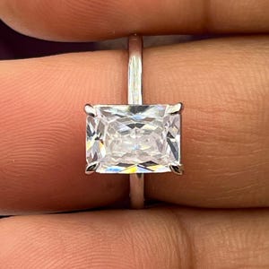 Può includere: Un anello d'argento con una grande pietra preziosa trasparente a taglio rettangolare. La pietra preziosa è incastonata con quattro griffe e la fascia è in argento lucido. L'anello è tenuto tra due dita.