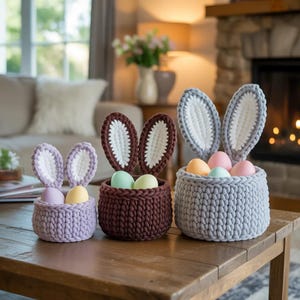 May include: Three handmade crochet Easter baskets in lavender, brown, and grey, each with bunny ears and filled with pastel-coloured eggs. The baskets are displayed on a wooden table, with a cosy living room setting in the background.