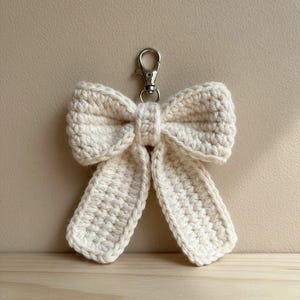 Puede incluir: Llavero de lazo tejido en color crema. El lazo está hecho de un hilo grueso, con un aspecto texturizado. Un cierre de metal plateado está unido en la parte superior para facilitar la fijación a llaves o bolsos. El lazo está centrado.