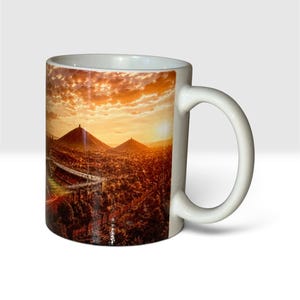 Op de afbeelding: Een witte keramische mok met een rondlopende fotoprint van een landschap bij zonsondergang. De afbeelding toont twee grote kegelvormige heuvels onder een heldere oranje en gele lucht, met een stadionstructuur op de voorgrond.