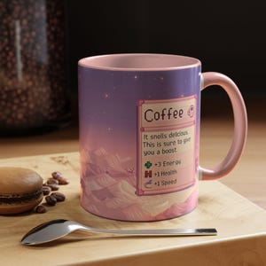 May include: A ceramic coffee mug with a pink handle and interior. The mug features a pixelated landscape design with text that reads "Coffee" and "It smells delicious. This is sure to give you a boost."