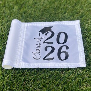 May include: White graduation banner with the text "Class of 2026" in black script and numbers, topped with a graduation cap graphic. The banner is rolled up at the end and is set on a bed of green grass.
