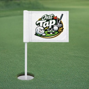 May include: A white golf flag displaying the text "Just Tap It" and a cartoon golf ball with an angry expression. The flag is on a white pole, set on a green golf course.