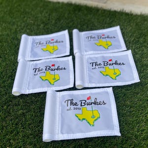 May include: Five white golf flags with the text "The Burkes est. 2013" and a yellow Texas-shaped graphic with a red flag. The flags are arranged on green grass.