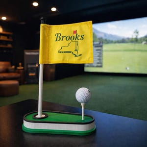 Puede incluir: Una maqueta de campo de golf con una bandera amarilla con el nombre "Brooks" y un contorno del estado de Nueva York. Una pelota de golf descansa sobre un tee. La pantalla está sobre una superficie negra, con un simulador de golf en el fondo.
