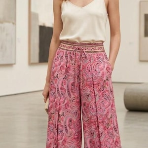 Pode incluir: Uma mulher usando uma camisola creme e calças palazzo rosa com estampa paisley. As calças têm cintura com cordão e pernas largas. O look é completado com sandálias douradas.