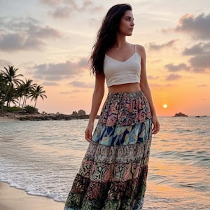 Puede incluir: Una persona con un top corto blanco y una falda larga escalonada con un colorido estampado de cachemira en una playa al atardecer. La falda presenta tonos de azul, rosa, verde y marrón. El cielo está lleno de colores cálidos.