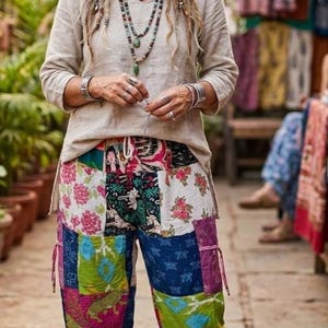 May include: A woman wearing a beige long-sleeve shirt and patchwork pants. The pants feature a variety of colorful floral and patterned fabrics. She is also wearing multiple necklaces and bracelets.