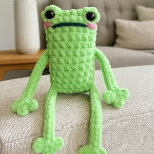 May include: A handmade, light green crochet frog plush toy. The frog has large black eyes, pink cheeks, and a dark green mouth. Its arms and legs are long and slender, with webbed feet. The plush is sitting on a neutral-colored surface.