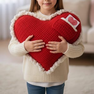 Peut inclure: Un grand coussin en crochet rouge en forme de cœur, avec une bordure blanche à volants. Une petite poche sur le coussin contient une carte avec une illustration de cœur. Le coussin est tenu.