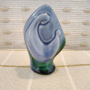 May include: A smooth, teardrop-shaped ceramic sculpture featuring a glossy glaze. The upper portion is a soft, muted blue, while the base transitions into a deep, rich forest green. The surface has a fluid, organic form with subtle curves.