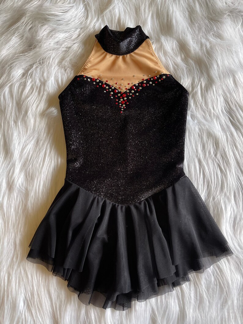 May include: Black figure skating dress with a sparkly bodice and a black tulle skirt. The bodice has a high neckline and a sheer panel with rhinestones and red accents.