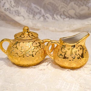 May include: A gold-colored tea set, including a lidded teapot and a creamer. The set has a textured, hammered appearance. The interior of the creamer is white. The set is displayed on a white lace surface.