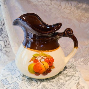 Op de afbeelding: Een vintage keramische kan met een bruine bovenkant en handvat, en een witte body. De kan heeft een fruitontwerp met rode kersen, gele peren en oranje fruit. De kan heeft een gebogen tuit en een decoratief handvat.