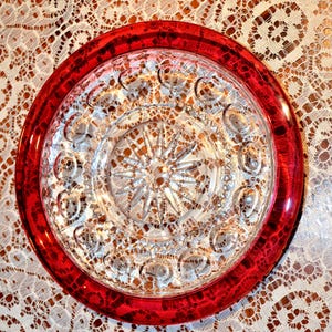 May include: A clear glass plate with a red rim. The plate has a detailed, starburst-like pattern in the center and a series of circular designs around the edge. The red rim has a marbled appearance. The plate is on a white lace tablecloth.