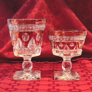 May include: Two clear, cut-glass goblets with square bases and ornate detailing. The glasses sit on a red patterned tablecloth, with a red cloth backdrop. The glasses are likely for serving beverages.