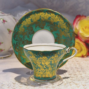 Könnte beinhalten: Ein türkisfarbenes Tee-Set mit goldenen Blumenmustern. Die Tasse hat einen weißen Innenraum und einen zarten Henkel. Die Untertasse befindet sich hinter der Tasse. Das Set steht auf einem transparenten Glasuntersetzer, mit einer weißen Tischdecke im Hintergrund.