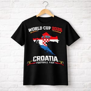 Puede incluir: Camiseta negra con un diseño gráfico de la bandera croata en forma de mapa. El texto "WORLD CUP 2026" está encima del mapa y "CROATIA FOOTBALL FAN" debajo. La camiseta está colgada de una percha de madera.