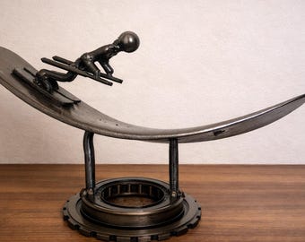 May include: A metal sculpture of a skier on a snowboard. The sculpture is made of dark metal and features a skier in a crouched position on a curved snowboard. The snowboard is supported by a gear-like base. The sculpture is a decorative item.
