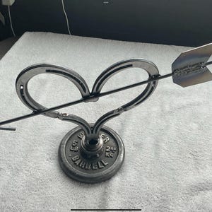 May include: A metal sculpture of a heart pierced by an arrow. The heart is formed from two horseshoes, and the arrow has a pointed tip. The sculpture is mounted on a weight plate base, with the text "5 LB BARBELL 2.25 KG".