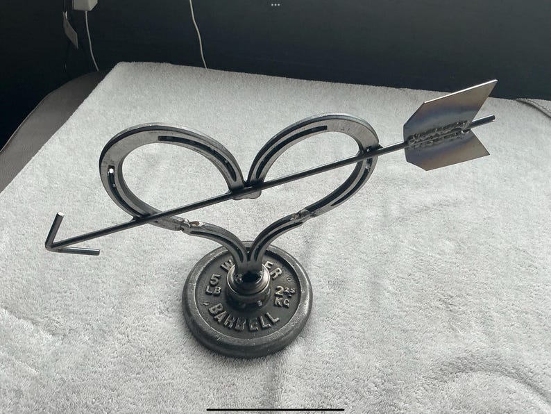 May include: A metal sculpture of a heart pierced by an arrow. The heart is formed from two horseshoes, and the arrow has a pointed tip. The sculpture is mounted on a weight plate base, with the text "5 LB BARBELL 2.25 KG".