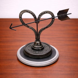 May include: A metal sculpture featuring a heart shape formed from two horseshoes, pierced by an arrow. The sculpture is mounted on a circular base with a dark gray top and a light gray rim. The arrow and heart are dark gray.